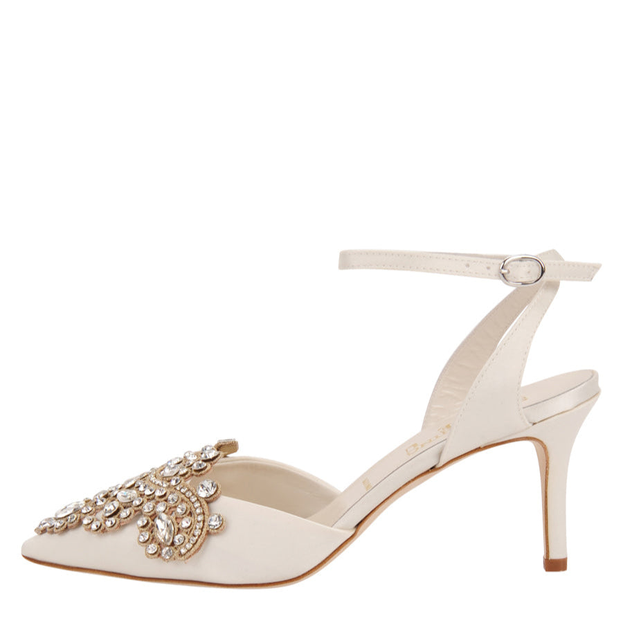 A white pointed toe pump with an ankle strap and jewel embellishments along the toe. The shoe has a sleek, elegant design and a stiletto heel.